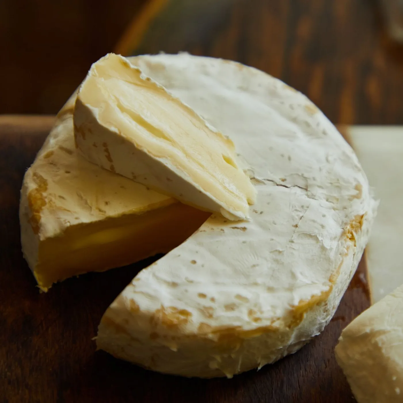 Peça de queijo brie sobre tábua de madeira, com uma cunha cortada mostrando a casca branca